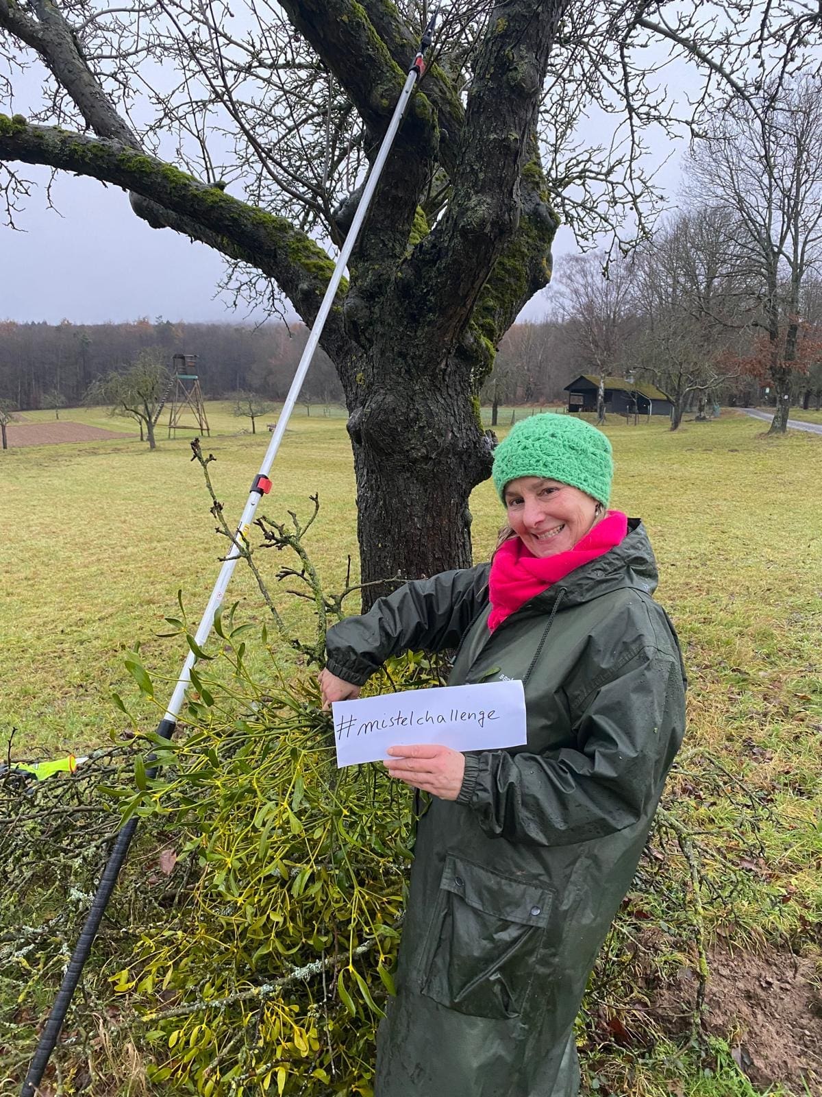 Frau mit Regenmantel und großer Mistel in rechter Hand unter einem Baum
